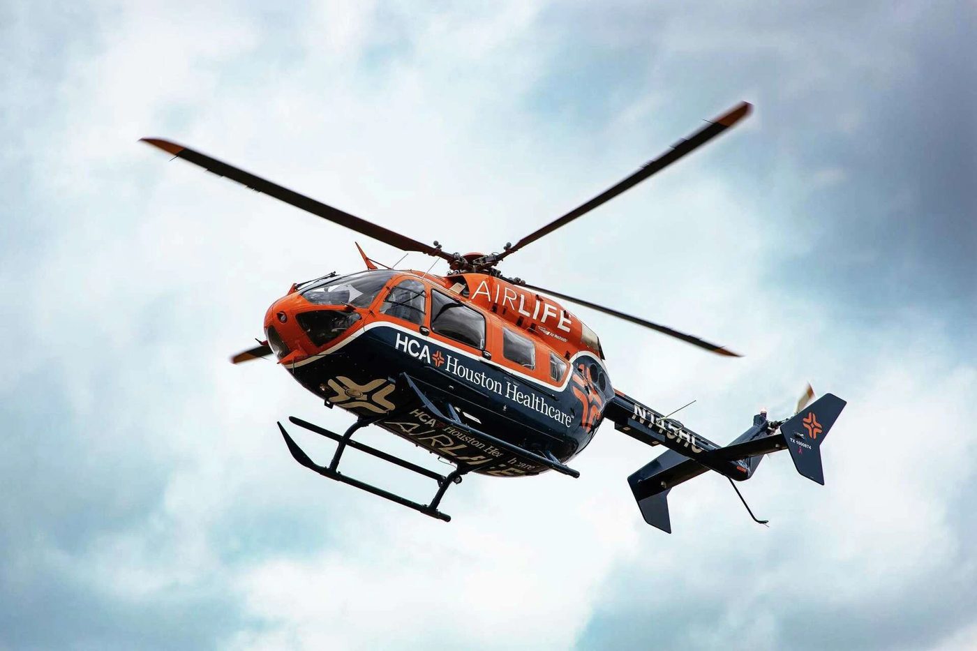 An HCA Houston Healthcare AirLife air ambulance helicopter in flight, seen from below against a bright cloudy sky.