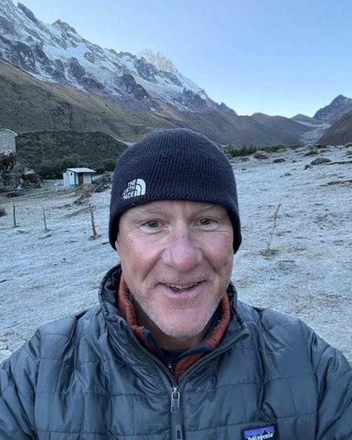 Dr. Rob Dickson in a beanie and insulated jacket on a frost-covered trail below snow-capped peaks.