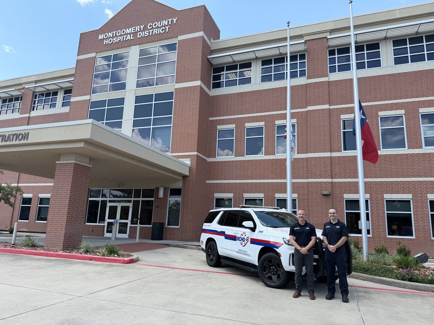MCHD Administration building with a white MCHD command SUV, two faculty members, and a Texas flag out front.