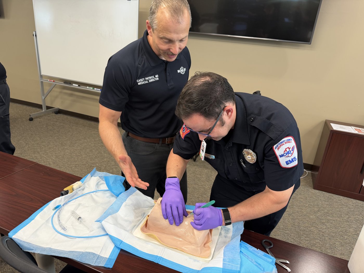 Dr. Casey Patrick in a navy MCHD polo supervising a medic performing a cricothyrotomy on a task trainer.