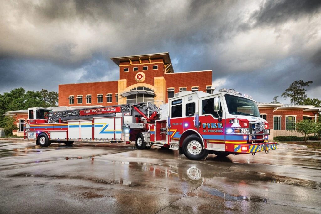 Two The Woodlands Fire Department apparatus — a ladder truck and an engine — parked on wet pavement outside a brick fire station under storm clouds.